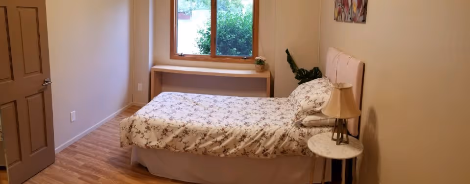 A small bedroom with a single bed covered in floral patterned bedding. Next to the bed is a round side table with a lamp on it. There is a window with a wooden frame letting in natural light, and a small shelf below the window with a small plant on it. The room has light-colored walls and wooden flooring.