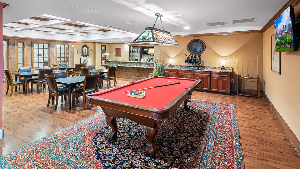 A spacious recreational room featuring a red felt pool table on a patterned rug in the center. Behind the pool table, there is a wooden cabinet with lamps and decorative items on top. To the left, there are several tables with chairs arranged for seating. The room has wooden flooring, beige walls, and a ceiling with recessed lighting and skylights. A flat-screen TV is mounted on the right wall.