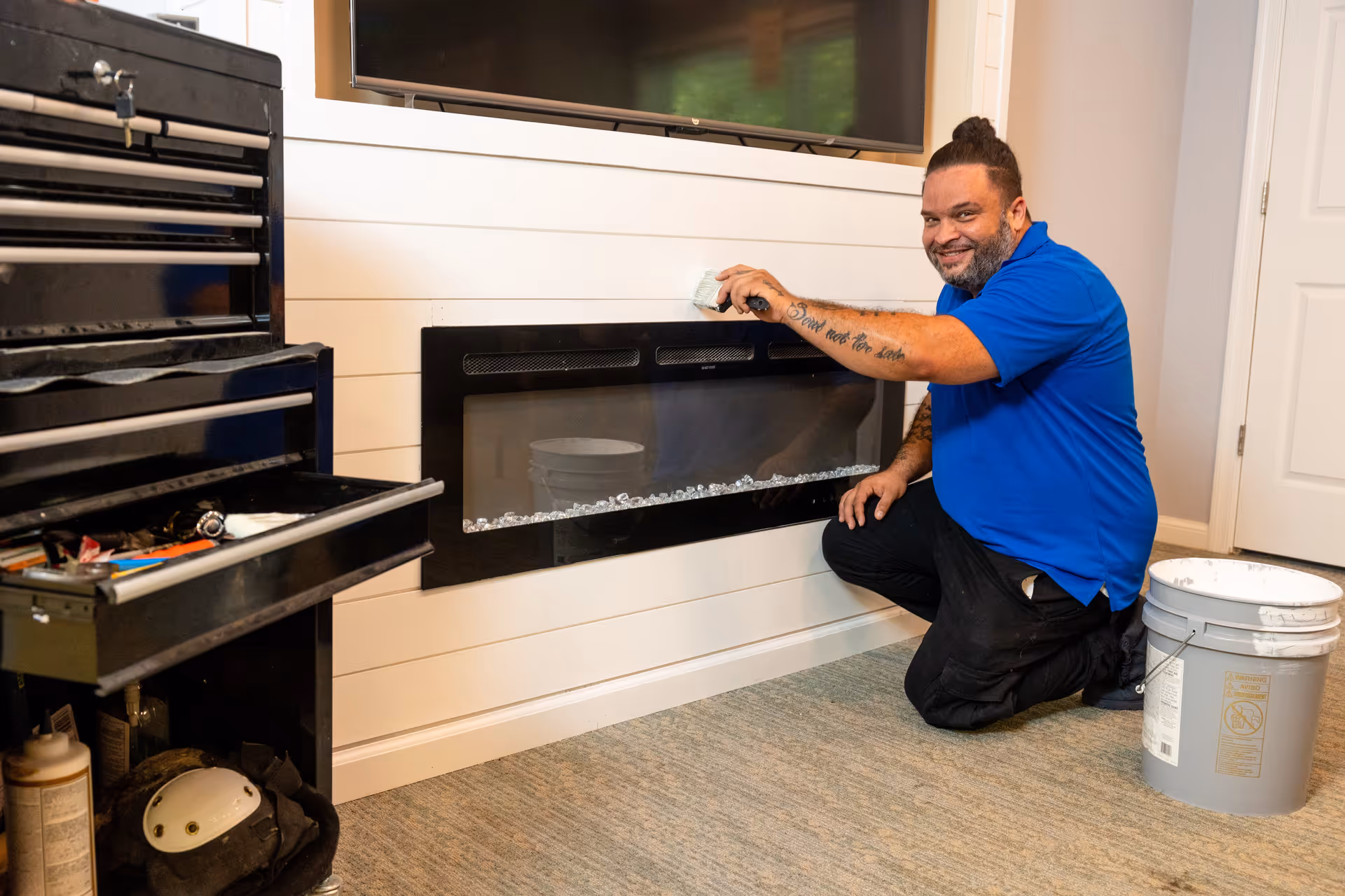 A man wearing a blue shirt and black pants is kneeling on the carpeted floor while painting white wooden paneling around a modern electric fireplace. There is a gray bucket with paint next to him and a black tool chest with open drawers on the left side. A flat-screen TV is mounted above the fireplace.