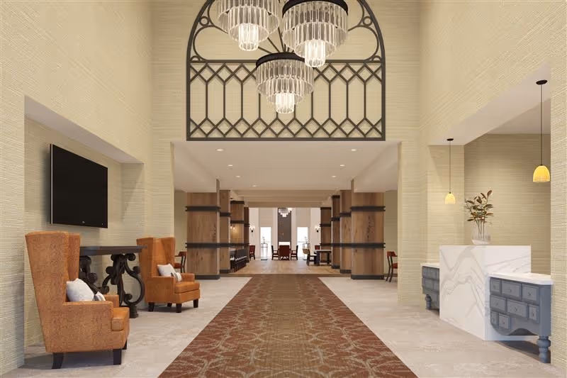 Spacious and elegant interior lobby area with high ceilings, a patterned carpet runner, two orange upholstered armchairs with pillows on the left, a mounted flat-screen TV above a decorative console table, and a reception desk with pendant lights and a vase with flowers on the right. The space features wooden columns with black metal accents and large hanging chandeliers.