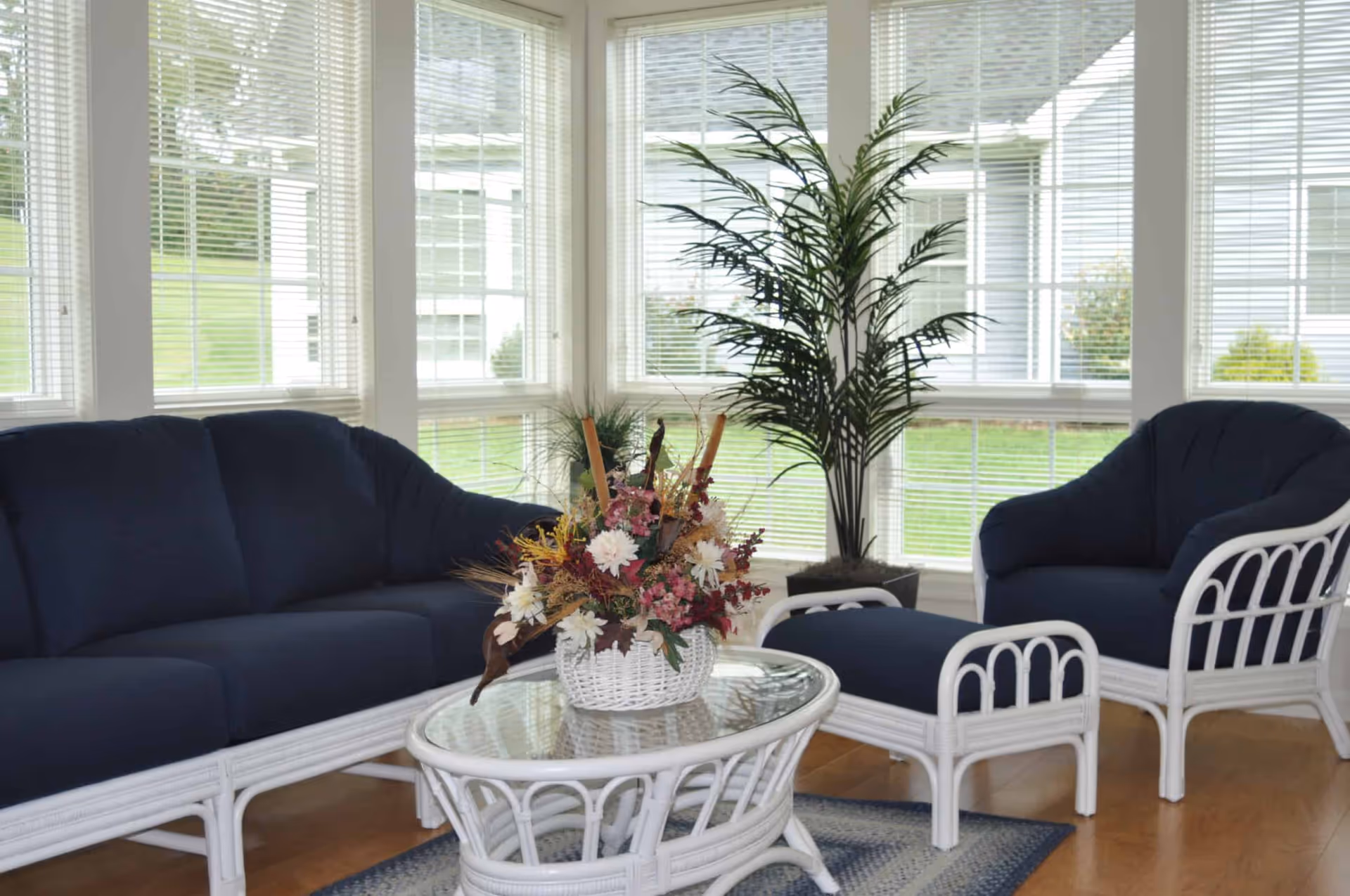 A bright living room area with large windows covered by white blinds. The room features a white wicker furniture set with dark blue cushions, including a sofa, an armchair, and an ottoman. A glass-top coffee table with a white wicker base holds a decorative basket of flowers. A tall green potted plant is placed near the windows, and a blue and white rug lies on the wooden floor.