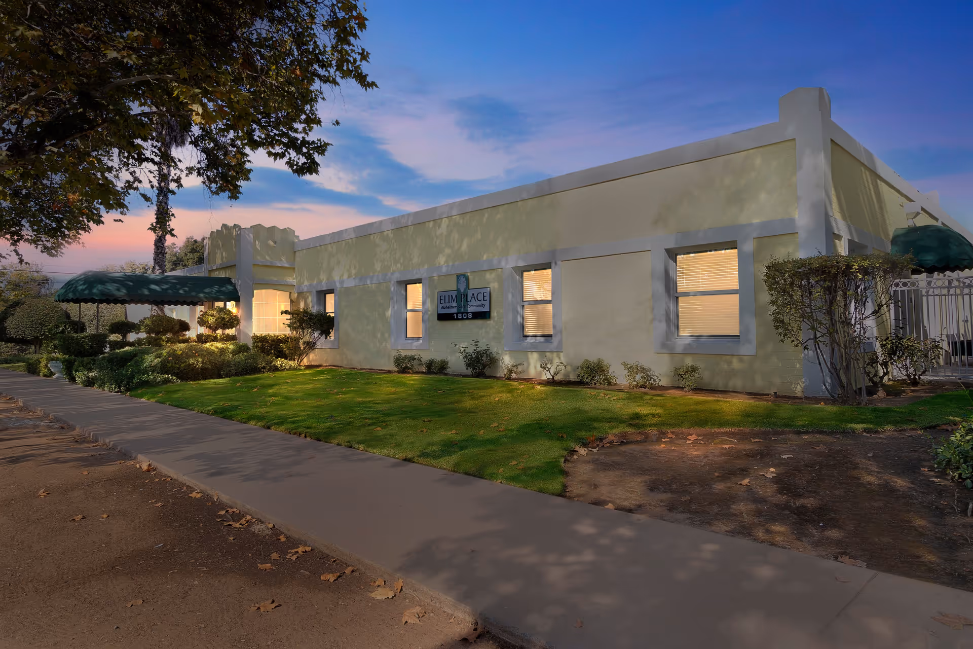 Exterior view of Elim's Place facility at dusk, showing a single-story building with light yellow walls and white trim. The building has several windows with blinds and a green awning over the entrance. There is a well-maintained lawn and landscaped bushes along the sidewalk in front of the building.