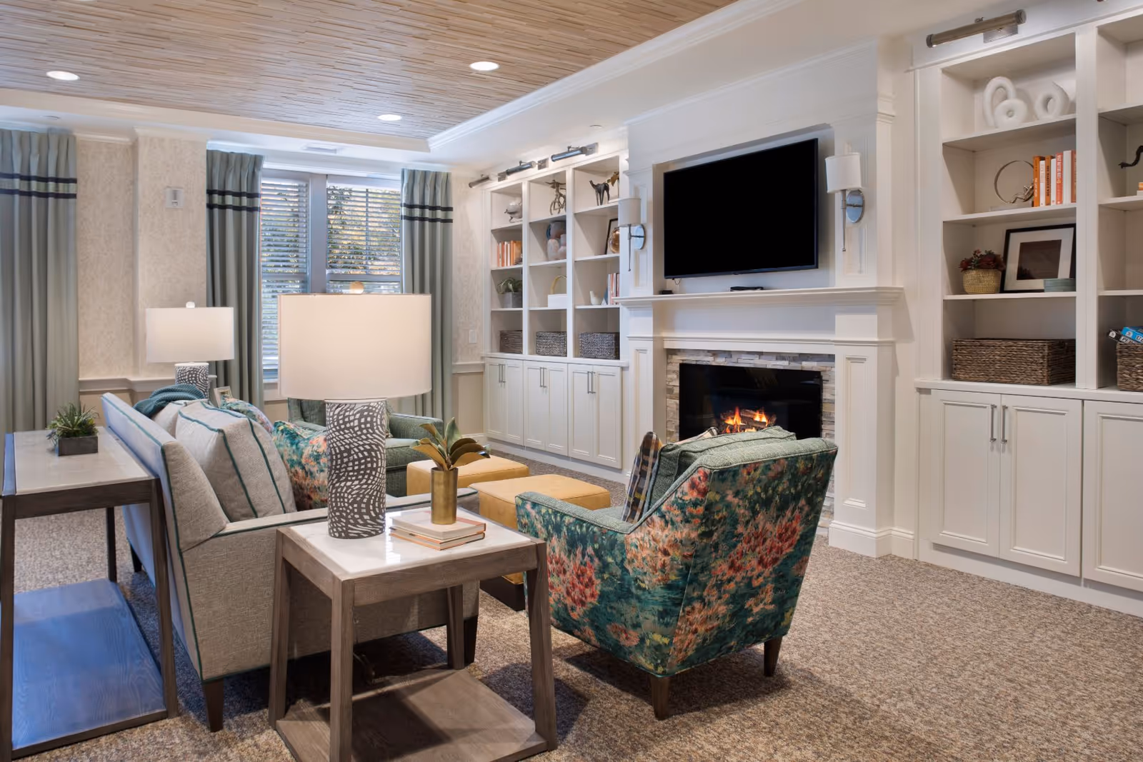 Cozy living room with a sofa, floral armchairs, side tables and lamps facing a fireplace and wall-mounted TV surrounded by built-in white shelving.