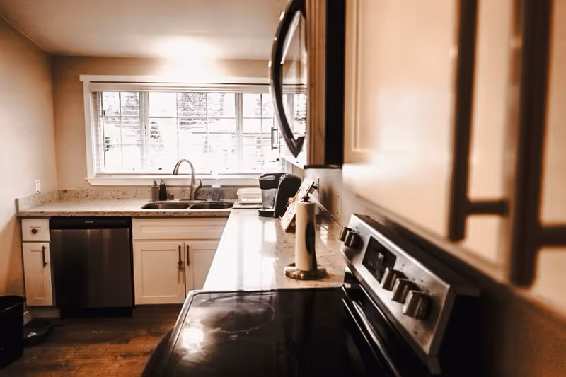 Bright kitchen with a stove and microwave on the right, a sink under a large window, and white cabinets and countertops.