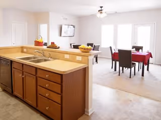 View of a kitchen area with a double sink and wooden cabinets in the foreground, and a dining area with two tables covered with red tablecloths and dark chairs in the background. The room is well-lit with natural light coming through large windows and a glass door.