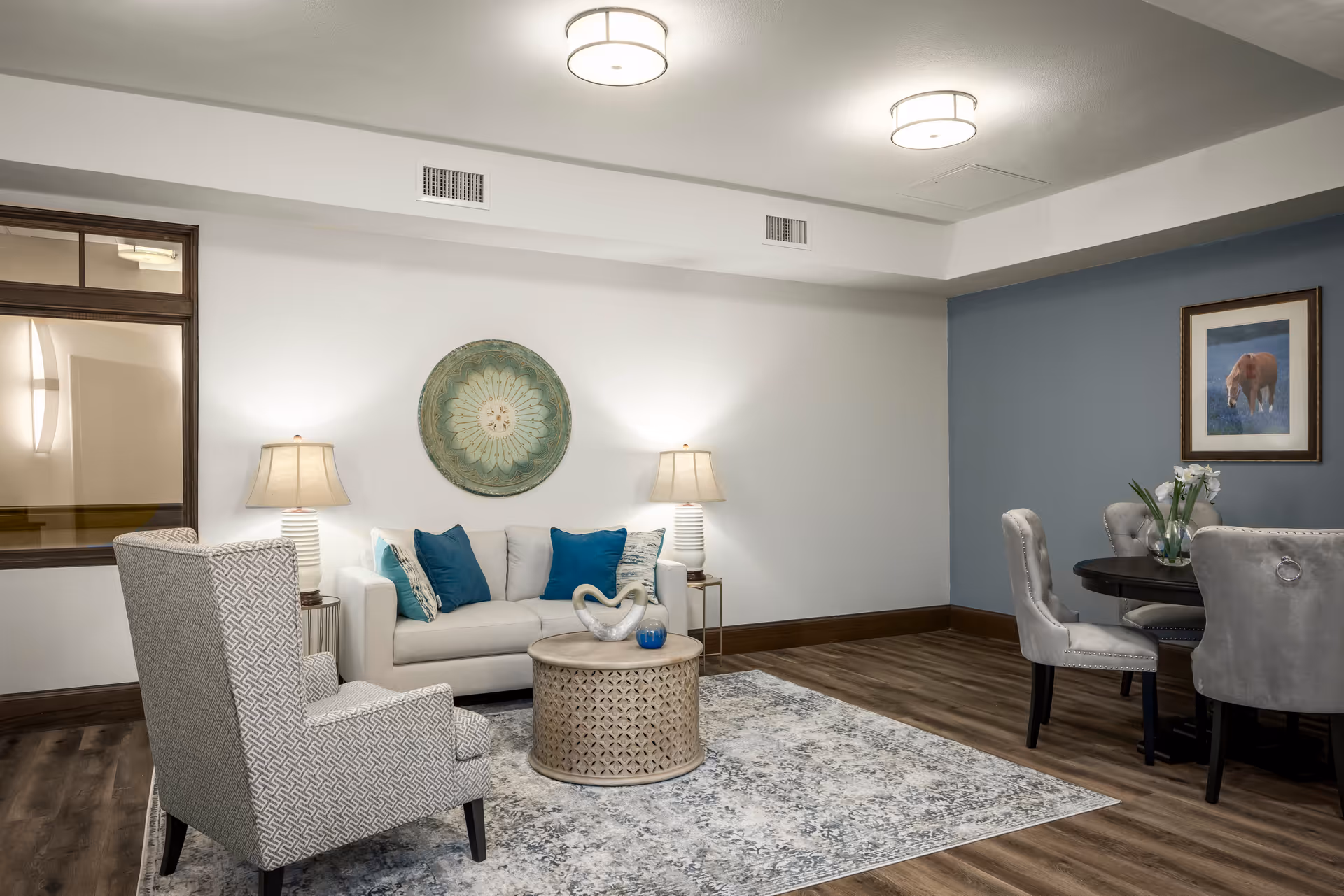 A cozy living room area with a white sofa adorned with blue and patterned pillows, two table lamps on either side, a patterned armchair, a round decorative coffee table, and a rug with a subtle design. In the background, there is a round dining table with four upholstered chairs, a framed picture of a horse on a blue accent wall, and a vase with flowers on the table.