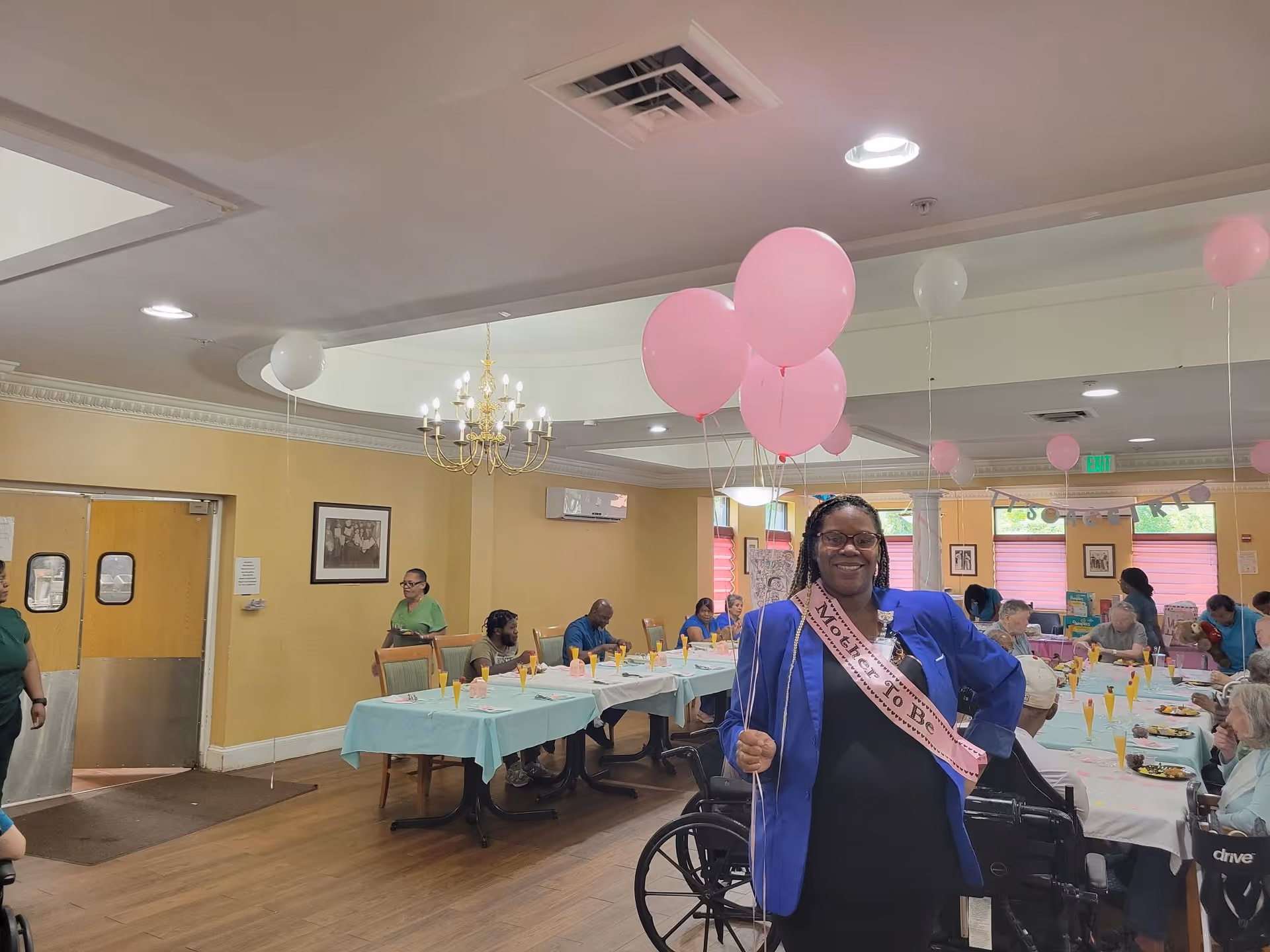 A smiling woman wearing a 'Mother To Be' sash and holding pink balloons stands in a decorated dining room where seniors sit at tables.