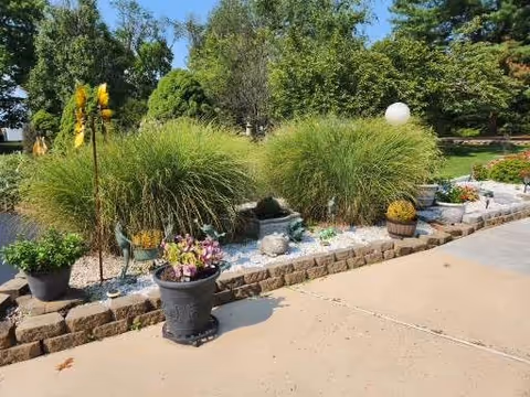 A landscaped outdoor garden area with various potted plants and ornamental grasses bordered by a low stone wall, adjacent to a paved walkway. Trees and greenery are visible in the background under a clear sky.