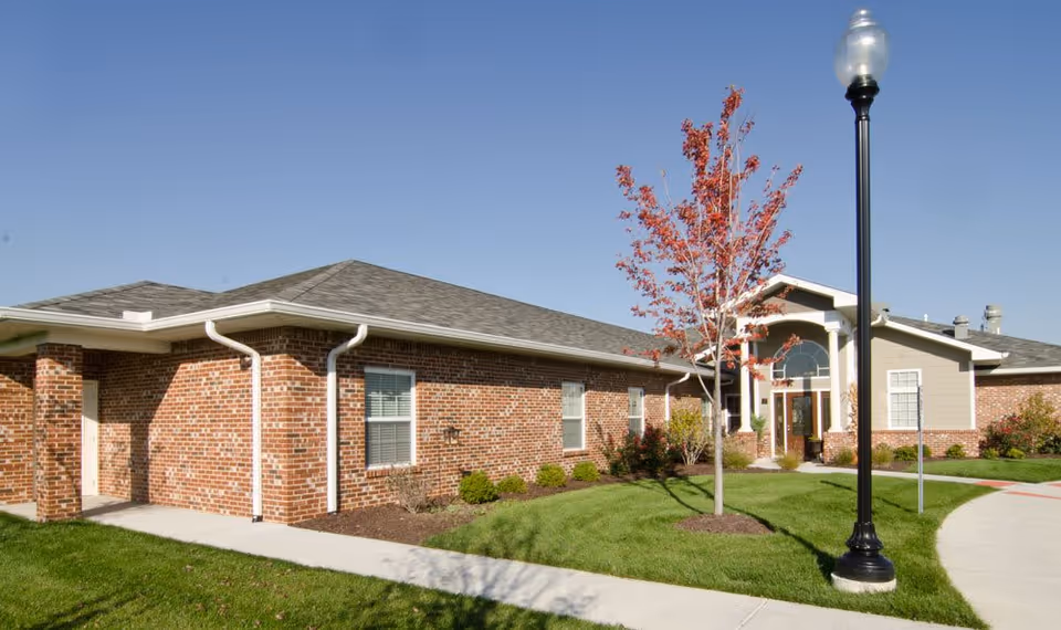 Exterior view of a single-story senior living facility building with red brick walls, a gray shingled roof, a small landscaped garden with a young tree with red leaves, and a black street lamp on a sunny day with clear blue sky.