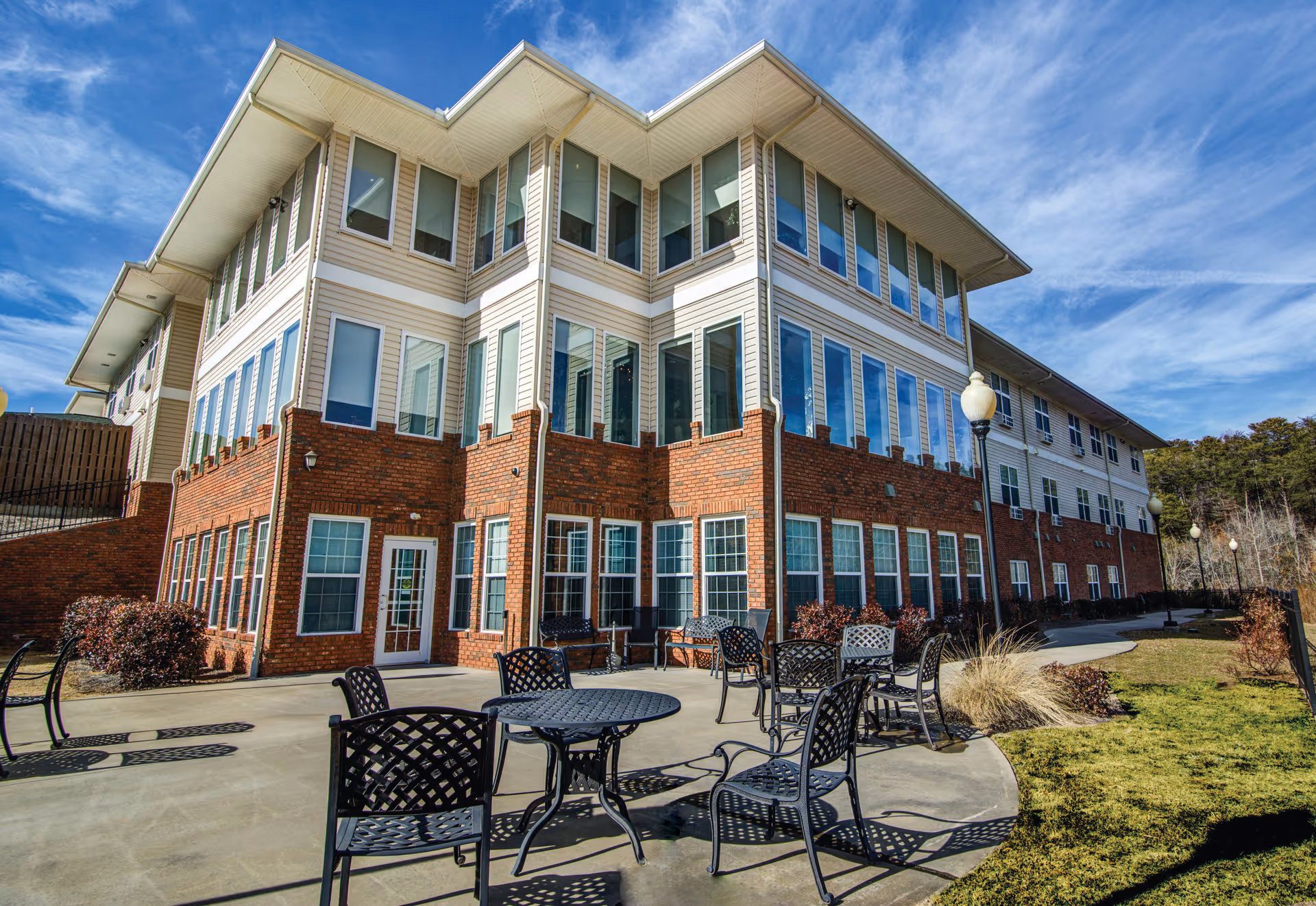 Exterior view of a multi-story brick-and-siding senior living building with a paved patio area and metal tables and chairs under a blue sky.