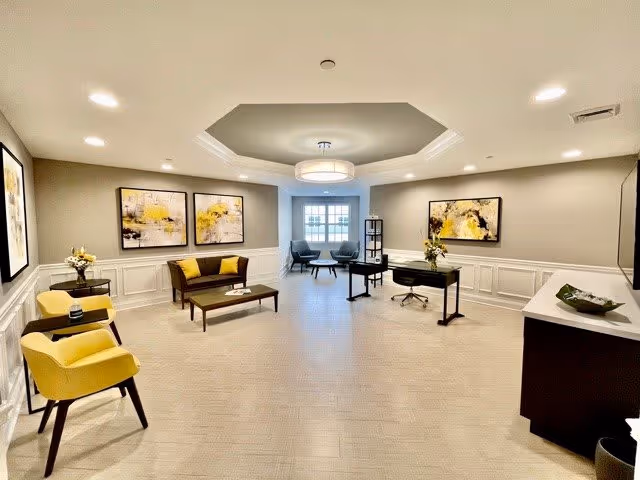 A spacious, well-lit interior room with beige walls and carpet. The room features modern furniture including a yellow armchair, a brown loveseat with yellow cushions, a coffee table, a desk with a chair, and two blue chairs near a window at the far end. The walls are decorated with abstract yellow and black paintings, and there are flower arrangements on side tables. The ceiling has a recessed design with a central light fixture.