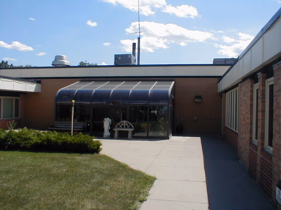 Brick single-story building with a glass-enclosed sunroom/entrance, walkway, benches and a small lawn in a courtyard.