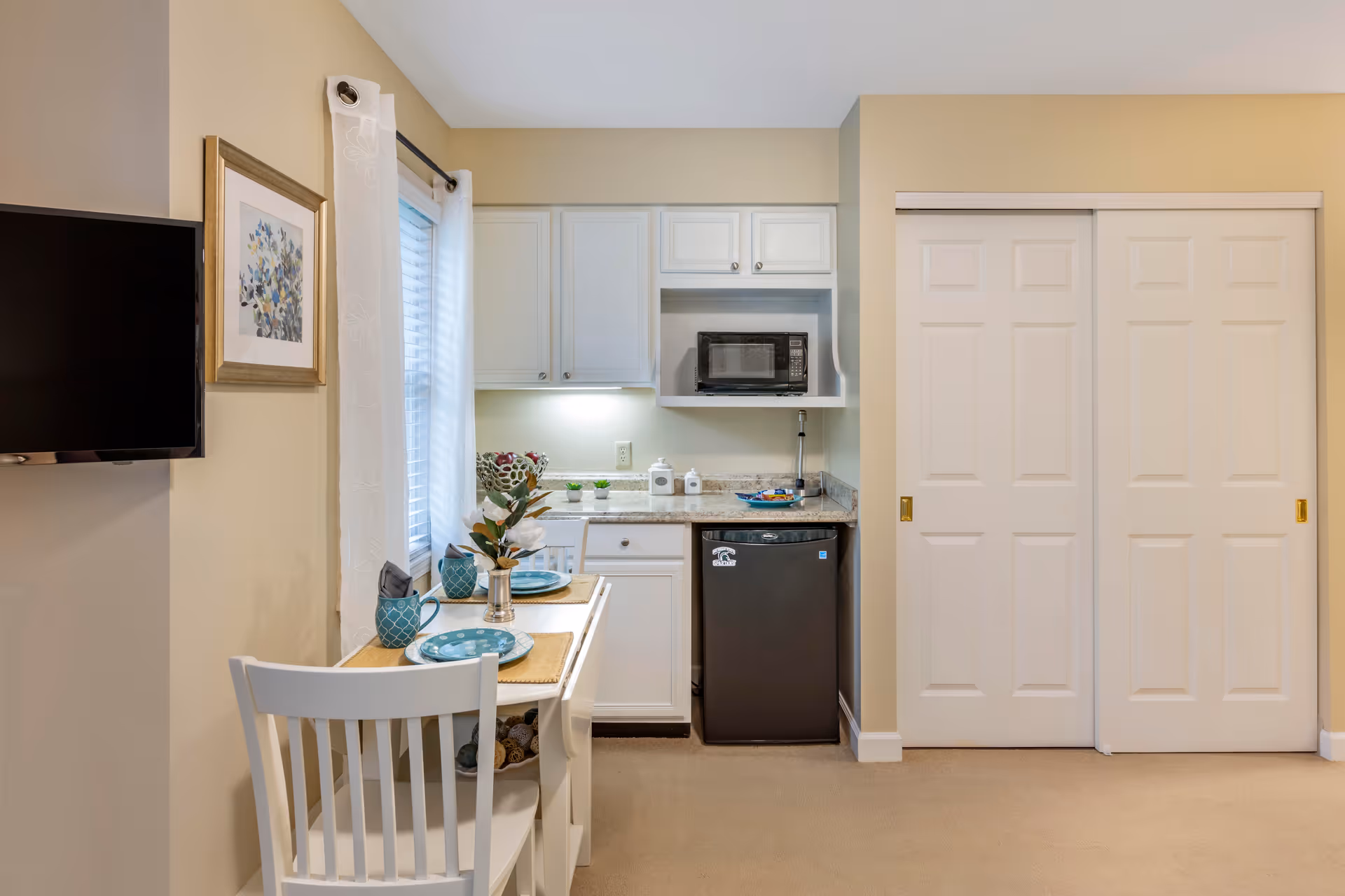 Small kitchenette area in an assisted living facility with white cabinets, a microwave, a mini refrigerator, and a small dining table set for two with blue plates and mugs. A flat-screen TV is mounted on the wall next to a window with white curtains, and there are sliding white closet doors on the right.