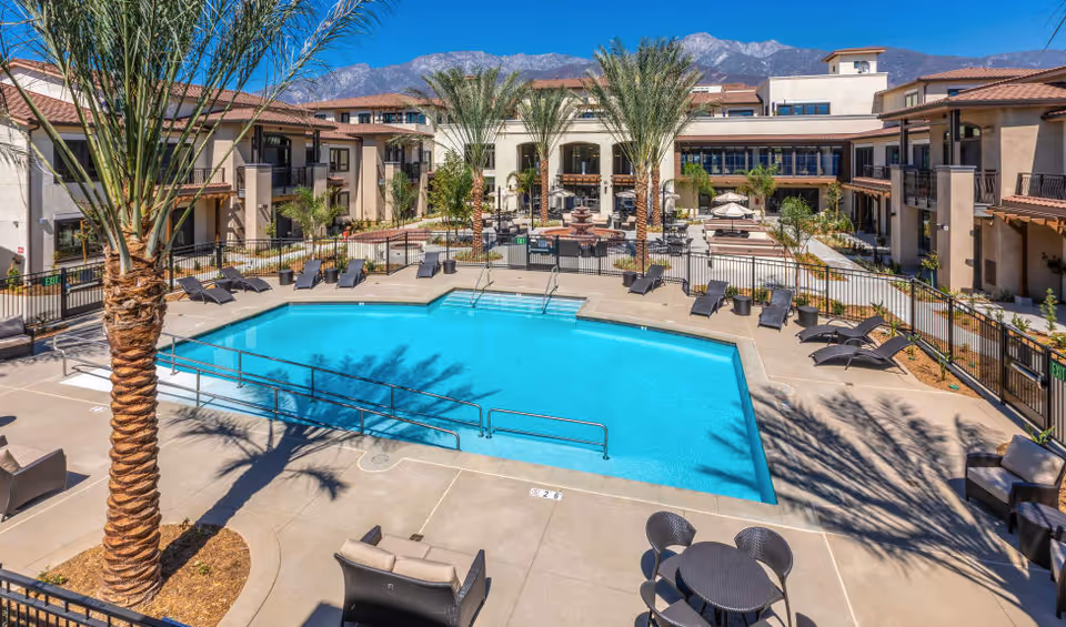 Outdoor swimming pool area at Merrill Gardens at Rancho Cucamonga with lounge chairs, palm trees, and a seating area. The pool is surrounded by a fence and the senior living facility building is visible in the background with mountains under a clear blue sky.