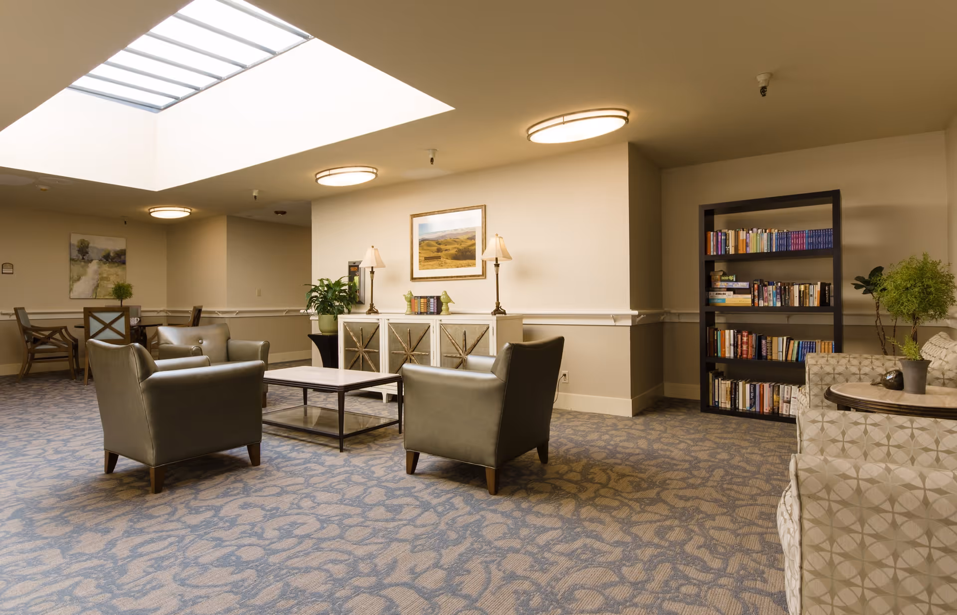 A well-lit common area with a skylight, featuring two leather armchairs around a coffee table, a bookshelf filled with books, a patterned sofa, and a sideboard with lamps and decorative items. The room has carpeted flooring and neutral-colored walls with framed artwork.