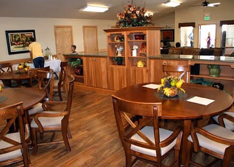 Interior view of a dining room with wooden round tables and chairs. There is a wooden cabinet with decorative items and flowers in the center. Two people are visible in the background near the walls and entrance door.
