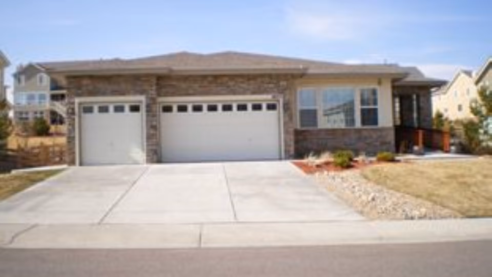 Front exterior view of a single-story house with a double garage and a driveway. The house has a stone facade and a small landscaped area with grass and shrubs.