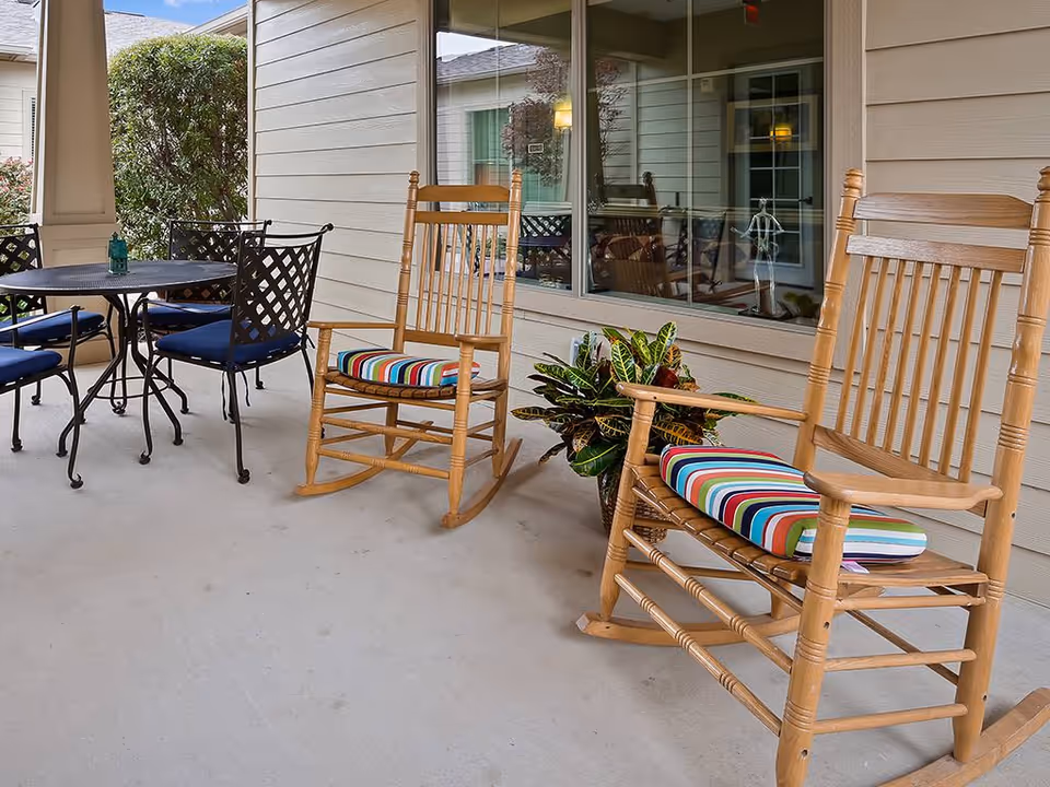 Covered outdoor patio area with two wooden rocking chairs featuring colorful striped cushions, a round black metal table with four matching chairs with blue cushions, and a potted plant near the window of a beige siding building.