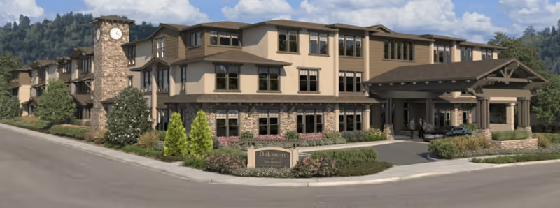 Exterior view of a large senior living facility building with a clock tower, multiple windows, and a covered entrance. The building is surrounded by landscaped greenery and a driveway with a car parked near the entrance. The sky is partly cloudy.
