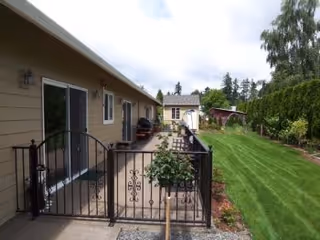 View of a residential outdoor area with a beige building on the left featuring sliding glass doors and a black metal gate. There is a concrete walkway and patio area with some potted plants. On the right side, there is a well-maintained green lawn bordered by shrubs and trees, extending towards another small building in the background under a partly cloudy sky.
