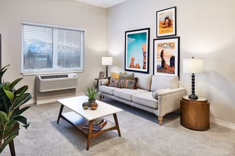 A bright and cozy living room with a beige sofa adorned with colorful pillows, a white coffee table with decorative plants, two side tables each with a lamp, a large window with blinds, and three framed art prints on the wall.