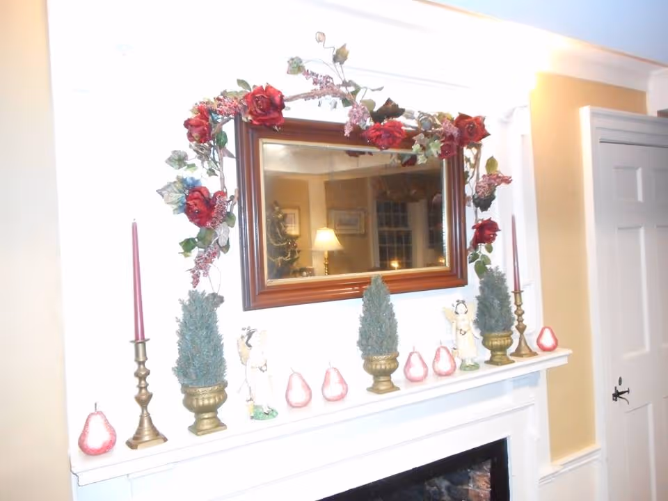 A decorated fireplace mantel with a large rectangular mirror framed with red flowers and green leaves above it. The mantel features two brass candle holders with pink candles, two small artificial evergreen trees in brass pots, small angel figurines, and several pink pear-shaped ornaments. The room has beige walls and a white door to the right.