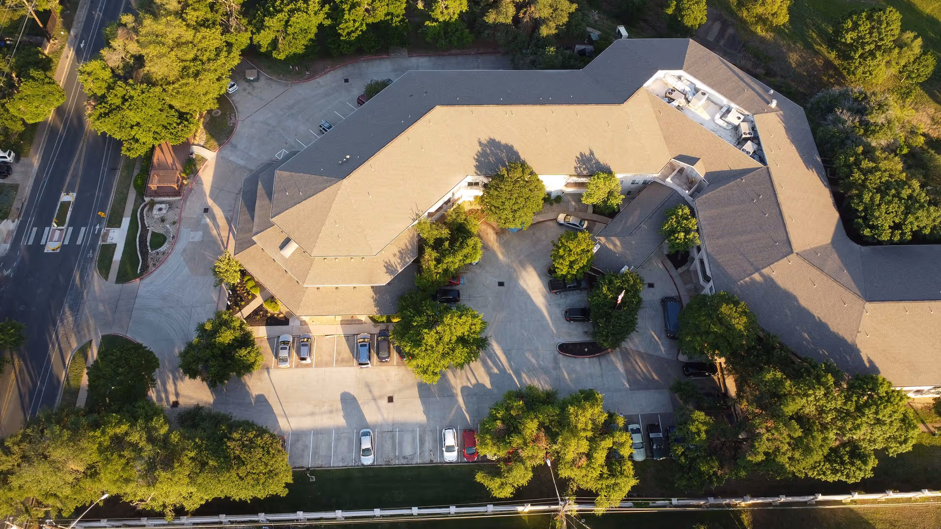 Aerial view of Solstice Senior Living at Austin showing a large building with a beige roof surrounded by trees and parking lots with several cars. The building is situated near a road with crosswalks and bike lanes, and there is greenery around the property.