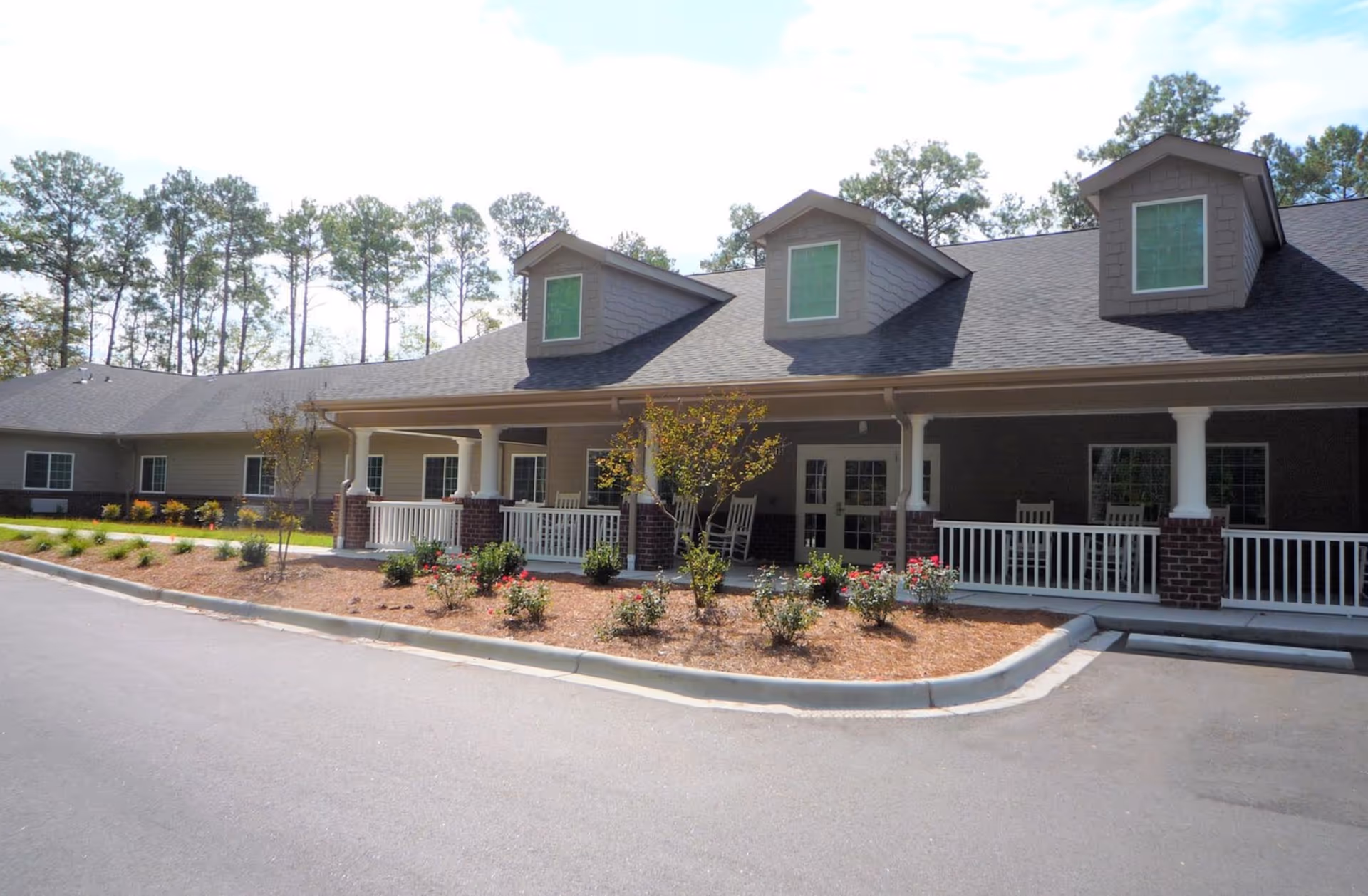 Front exterior of a single-story senior living building with a covered porch, rocking chairs, and landscaped flower beds.