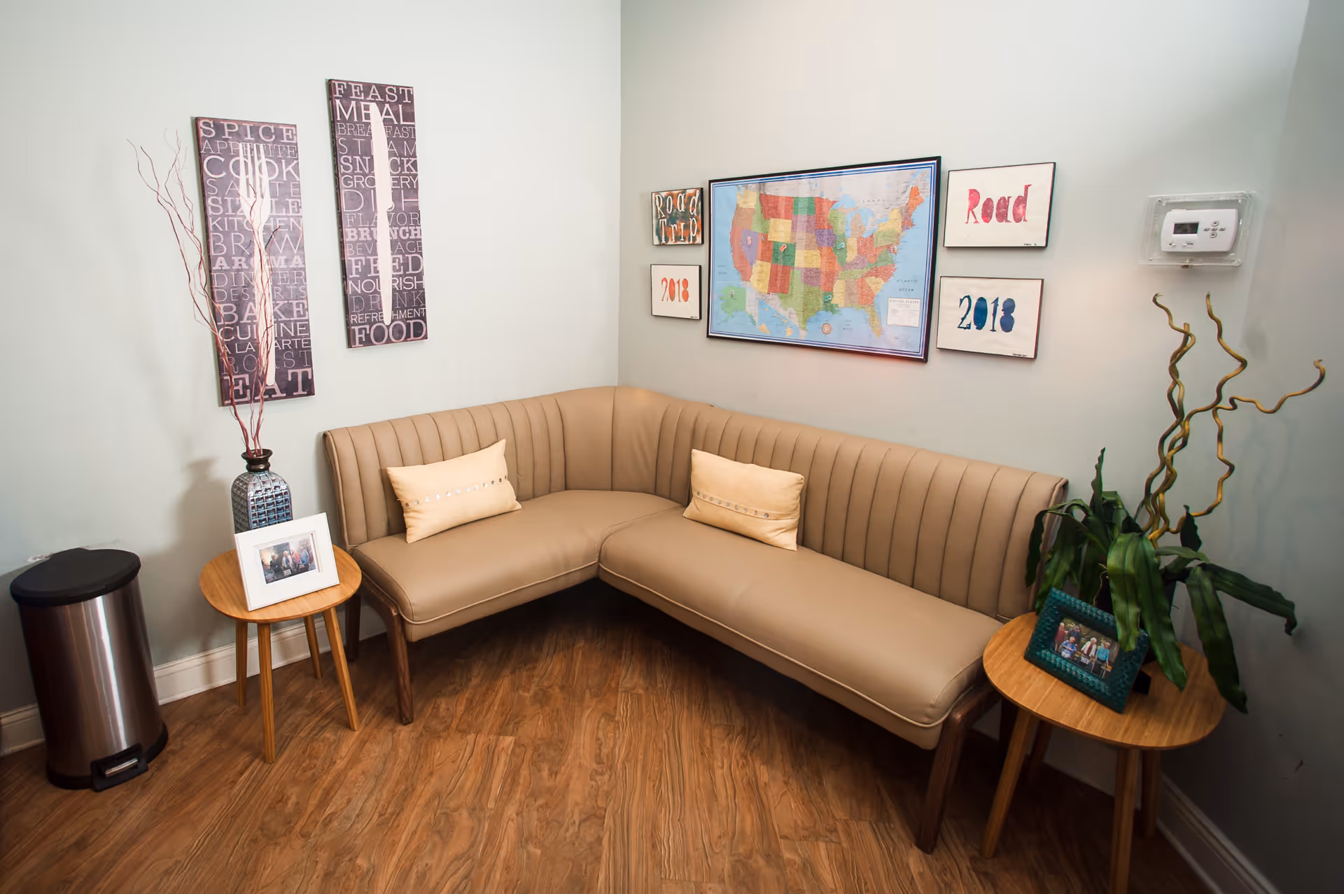 Corner seating area with a tan upholstered L-shaped bench, two small wooden side tables, wall art including a framed U.S. map, and potted plants.
