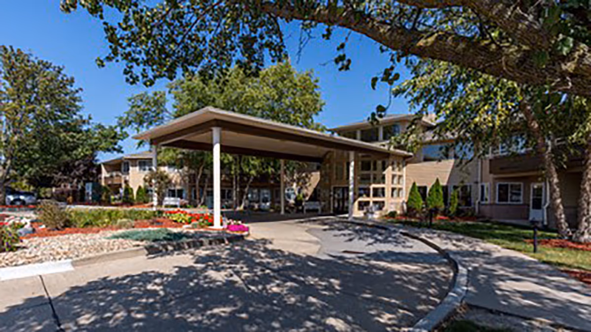 Front entrance of a senior living building with a covered porte-cochere, landscaped flower beds and trees.