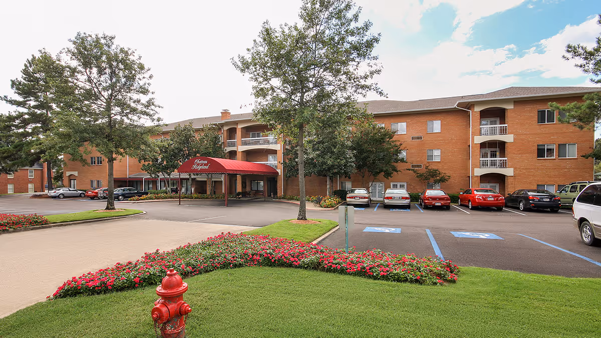 Exterior view of Holiday Chateau Ridgeland, a three-story brick building with a red canopy entrance. There are several trees, a flower bed with red flowers, a red fire hydrant, and a parking lot with cars and handicapped parking spaces.