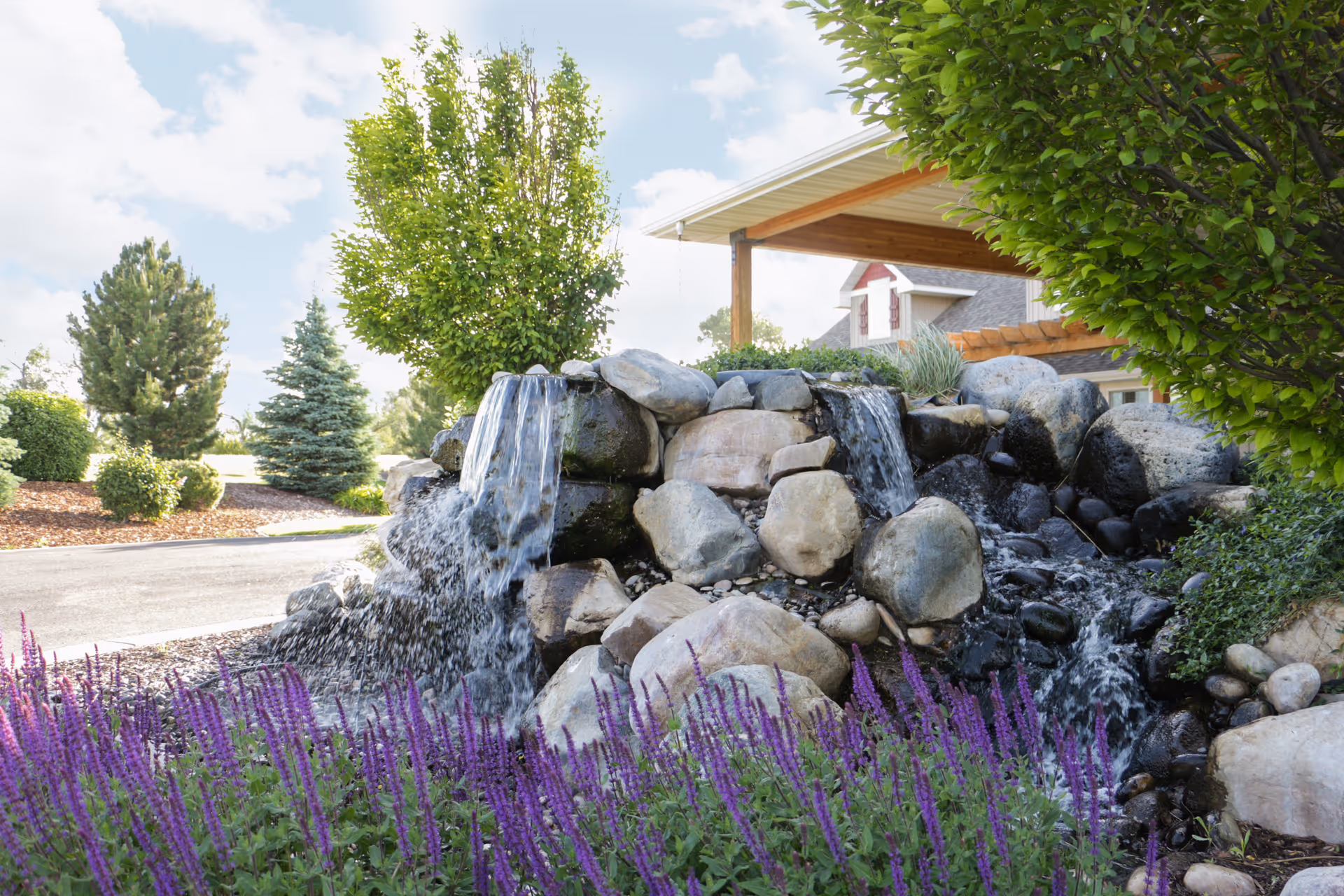 A landscaped outdoor area with a rock waterfall, purple flowering plants, and trees near a covered building entrance.