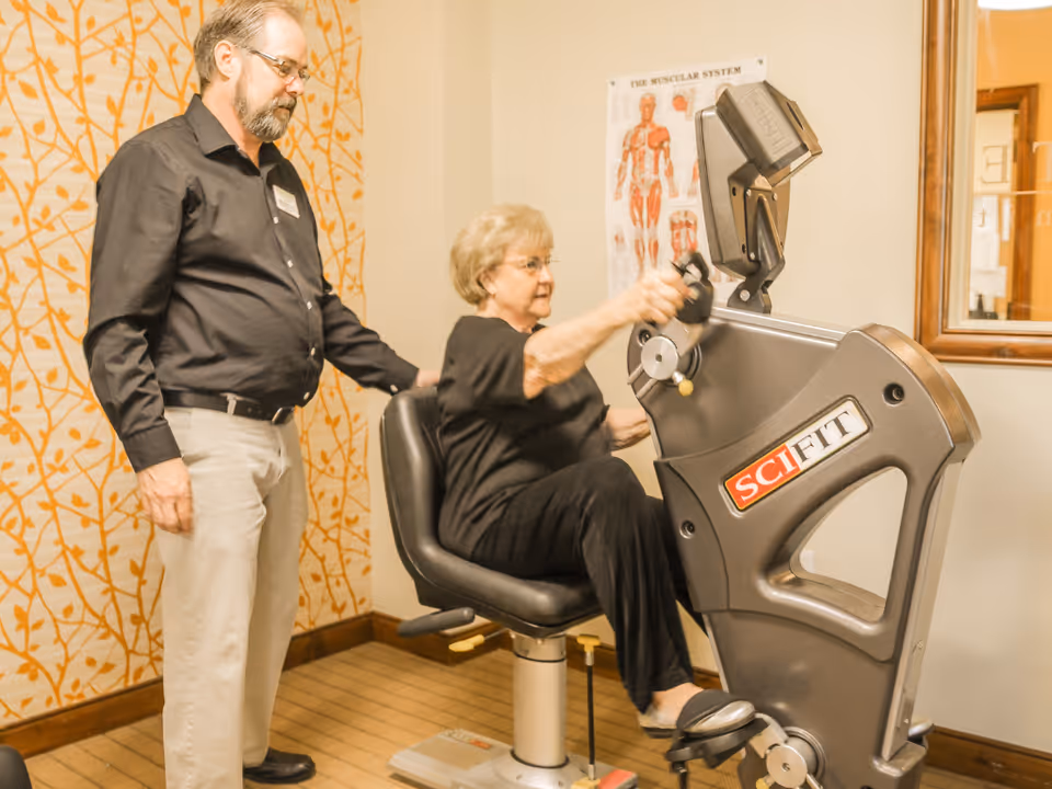 An elderly woman using a SciFit exercise machine while a man stands beside her offering support in a room with patterned wallpaper and a muscular system poster on the wall.