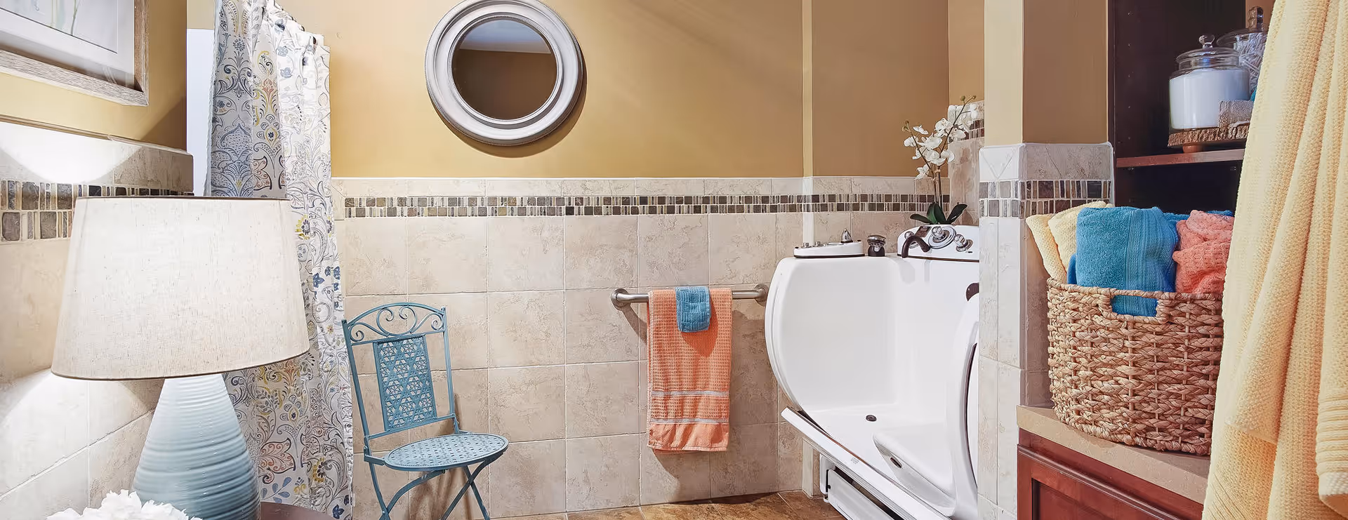 A well-lit bathroom featuring a walk-in bathtub with a door, a blue metal chair, a towel rack with orange and blue towels, a round mirror on the wall, a lamp on a small table, and a shelf with a basket of colorful towels and jars.