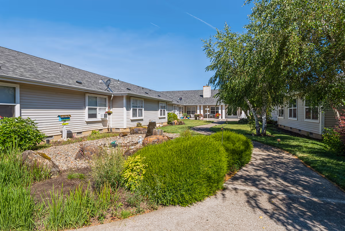Sunlit landscaped courtyard with a paved walkway between single-story residential buildings and trees.