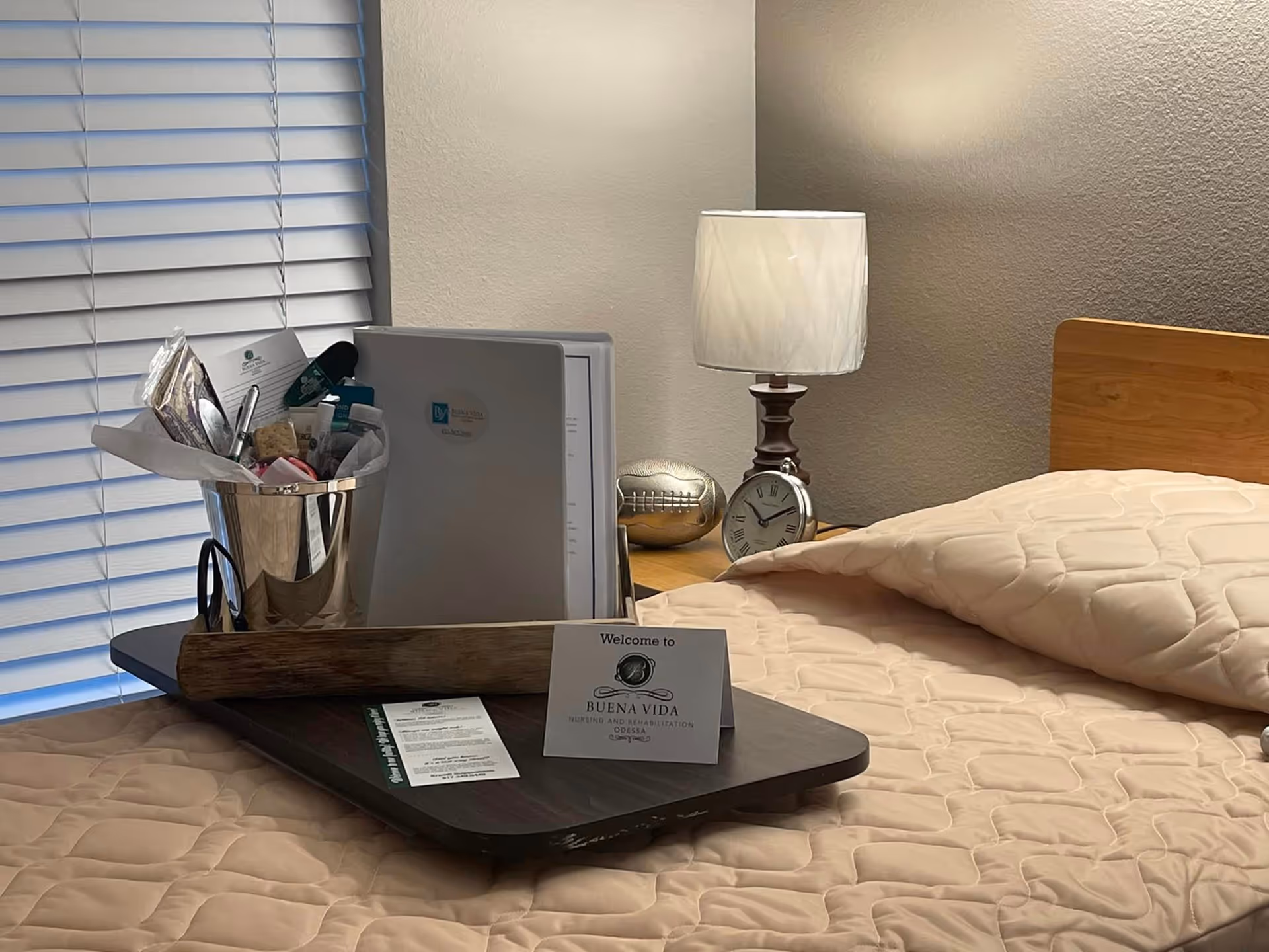 A neatly made bedroom bed with a wooden headboard, a tray holding a welcome card, toiletries, and paperwork, and a bedside table with a lamp and clock.