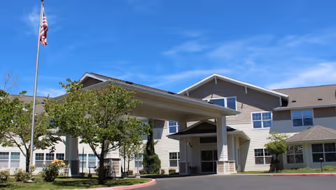 Exterior view of a senior living facility named Mallard Landing by Cogir, showing a two-story building with beige siding, a covered entrance, several windows, a flagpole with an American flag, and landscaped greenery under a clear blue sky.