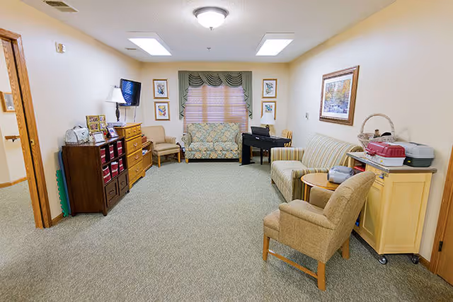A cozy living room area in a senior living facility with beige walls and carpeted floor. The room features a floral-patterned loveseat under a window with green valances, a striped sofa, and two beige armchairs. There is a small piano, a wooden cabinet with storage bins, a TV mounted on the wall, and framed artwork on the walls. Various small items and a basket are placed on the furniture.