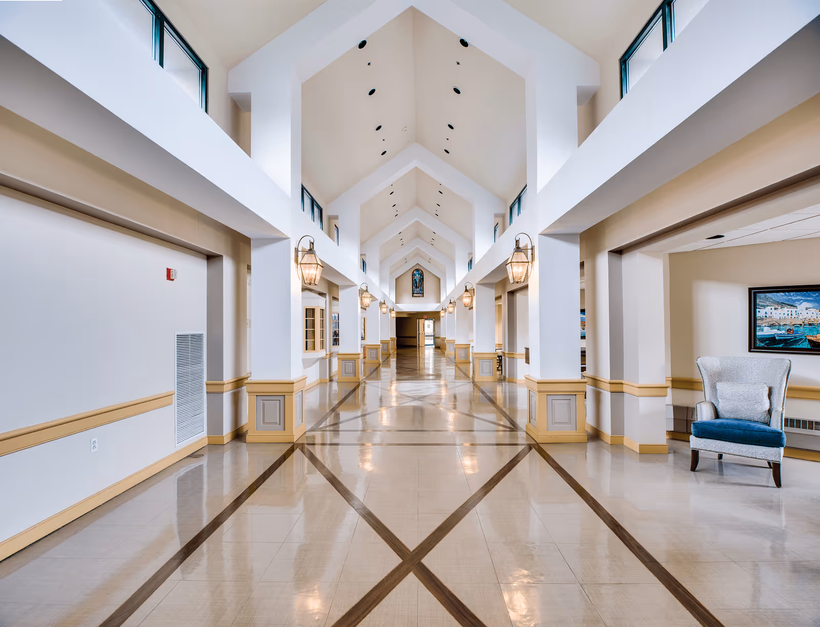 A long, bright hallway with a high vaulted ceiling featuring multiple triangular arches. The floor has a polished tile design with dark diagonal lines forming a pattern. The walls are painted white with beige wainscoting and decorative columns. Lantern-style wall lights line the hallway, and there is a blue upholstered armchair with a pillow on the right side near a framed painting. Windows near the ceiling allow natural light to enter.