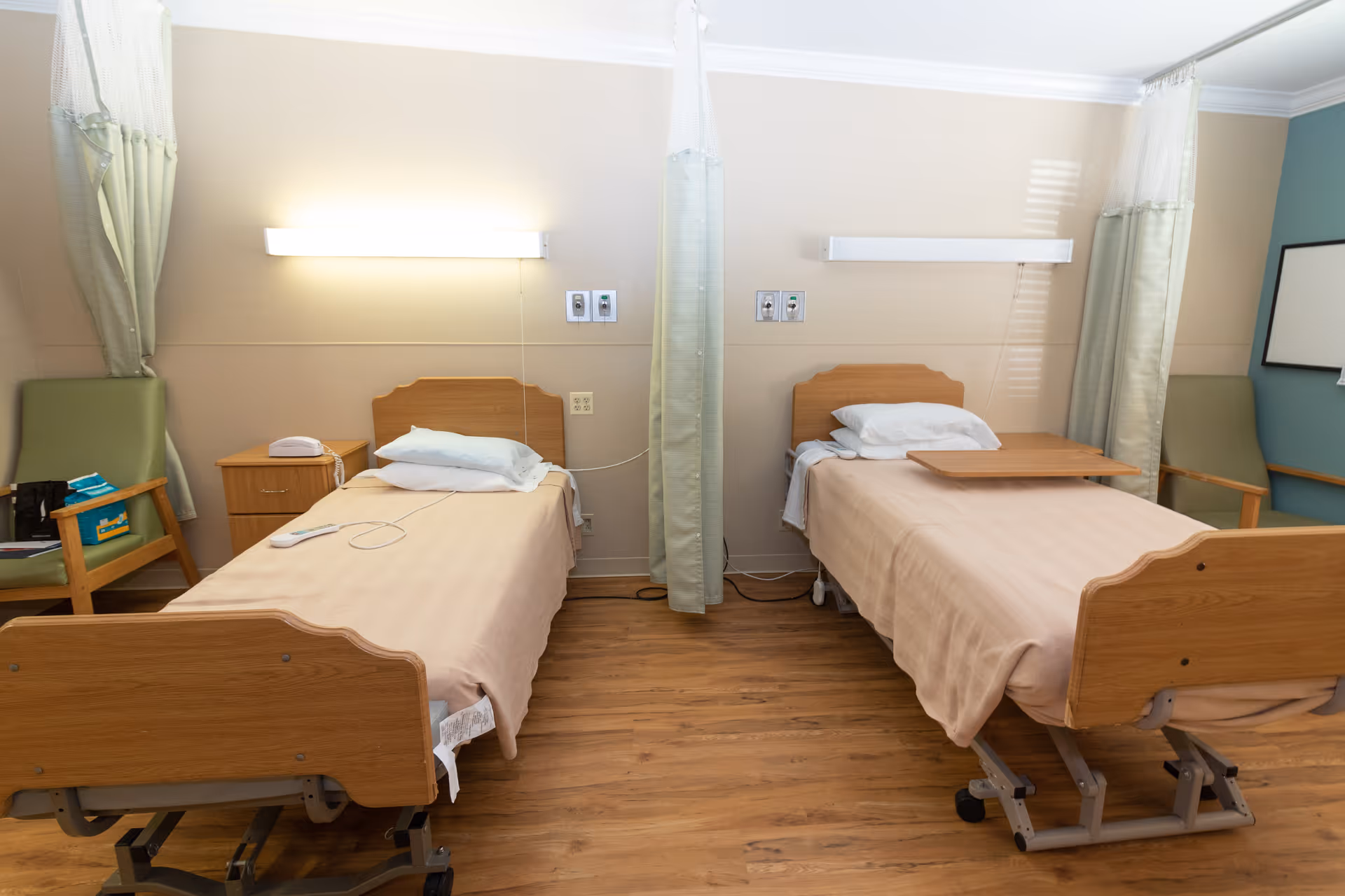 A room with two hospital-style beds separated by a privacy curtain. Each bed has a wooden headboard and beige bedding. There are green chairs next to each bed, a small wooden nightstand with a telephone, and wall-mounted lights above each bed. The floor is wooden, and the walls are painted in neutral tones.