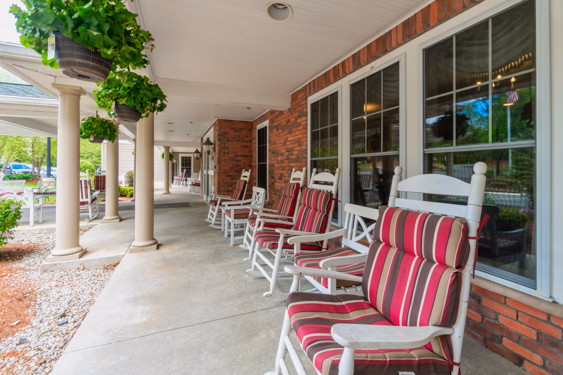 Covered front porch with white rocking chairs featuring red-striped cushions, hanging plants, columns, and a brick exterior wall.