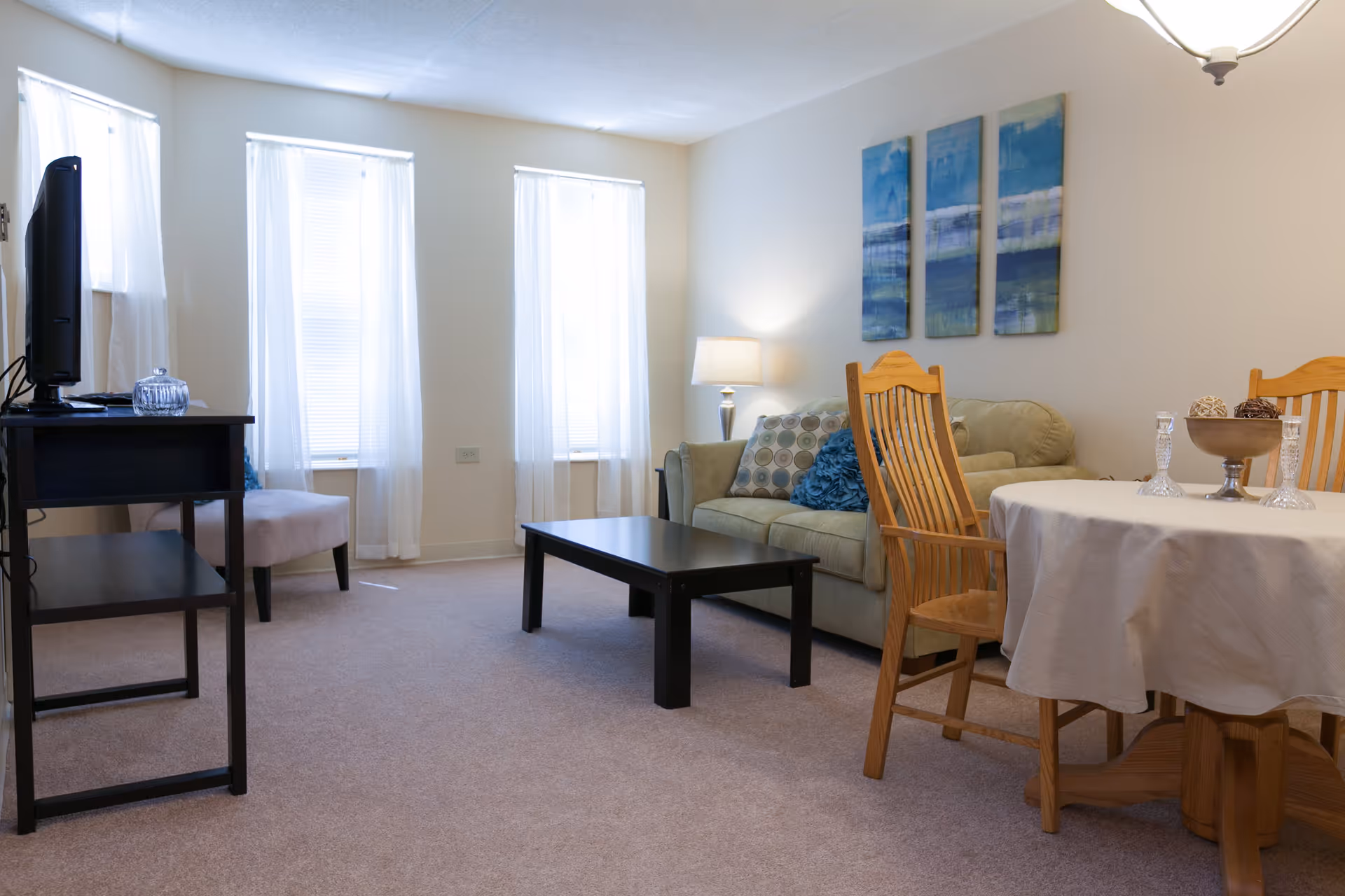A cozy living room with beige carpet and walls, featuring a light green sofa with decorative pillows, a black coffee table, a small black TV stand with a television, a beige upholstered chair, and a round wooden dining table with a white tablecloth and wooden chairs. Three blue abstract paintings hang on the wall above the sofa, and a table lamp provides soft lighting.