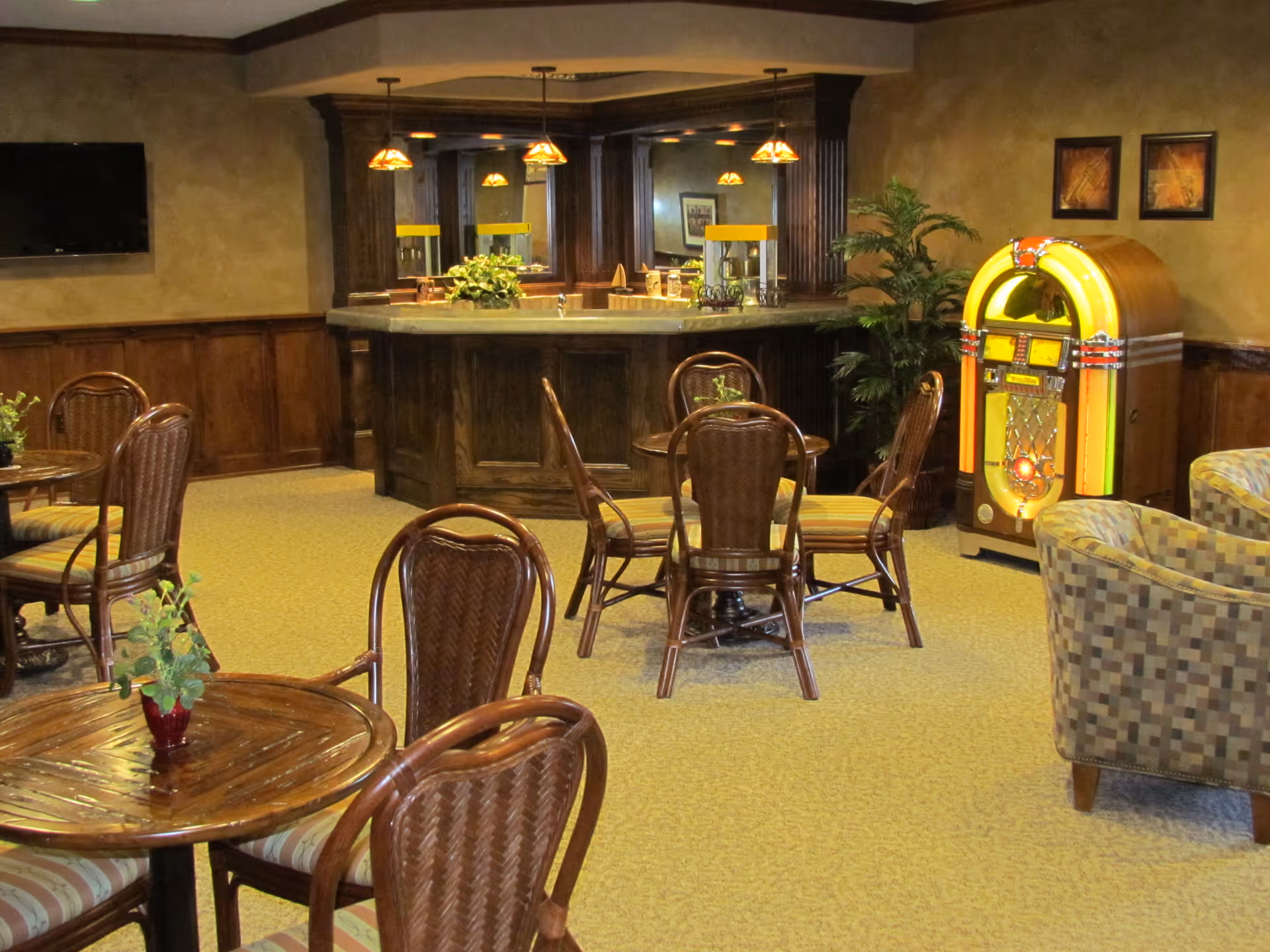 A cozy common area with wooden tables and chairs, a small bar with hanging pendant lights, a jukebox, a potted plant, and a wall-mounted TV. The room has warm lighting and carpeted floors.