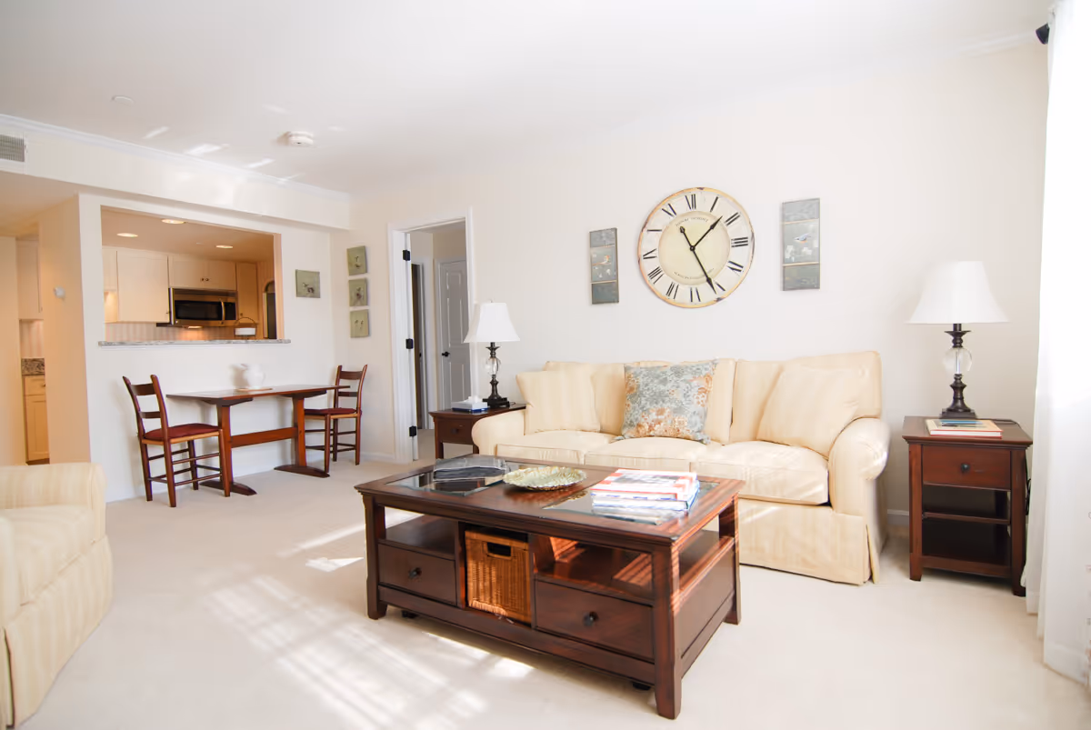 Bright living room with a beige sofa, wooden coffee table, side tables with lamps, and a small dining area near an open kitchen.