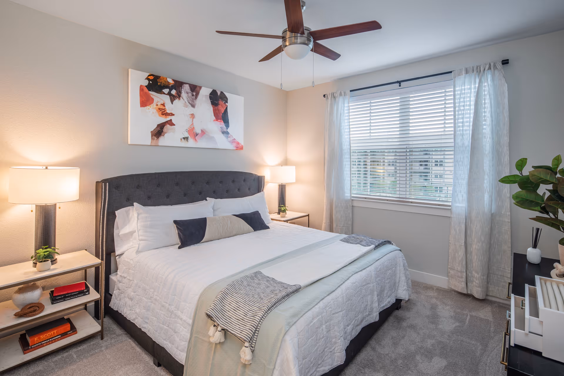 A neatly made bed with white bedding and a decorative pillow in a bedroom. Two bedside tables with lamps and small plants flank the bed. A large window with white blinds and light curtains lets in natural light. A ceiling fan with wooden blades is mounted on the ceiling. There is a piece of abstract art hanging above the bed and a dresser with decorative items on the right side of the room.