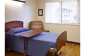 A simple bedroom with a single hospital-style bed covered with a blue blanket, a brown cushioned chair, a small framed picture on the wall, and a window with vertical blinds letting in natural light.