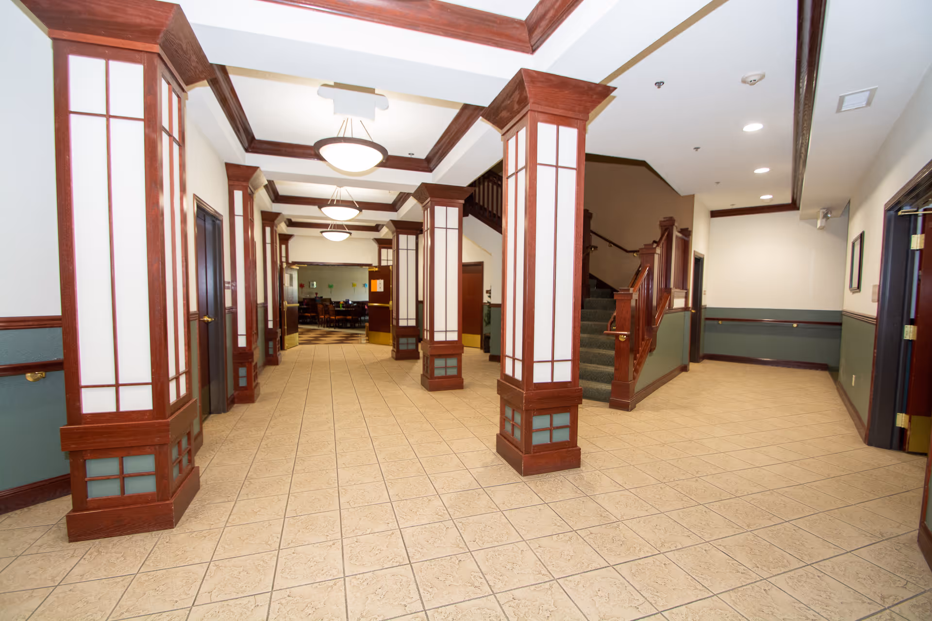Interior hallway of a senior living facility with beige tiled floor, wooden columns with white panels, green and white walls, ceiling lights, and a staircase leading to an upper floor. Doors line the hallway and a dining area is visible in the background.