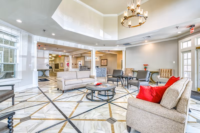 A bright and spacious senior living facility common area with marble patterned tile flooring, beige sofas with red cushions, several chairs, a round coffee table with magazines, and a chandelier hanging from a tray ceiling. Large windows and glass doors allow natural light to fill the room.