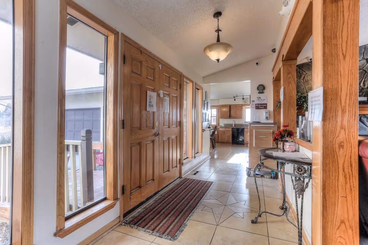 Interior view of a senior living facility entrance area with a wooden double door, large windows on the left, a striped rug on the tiled floor, and a small decorative table with flowers on the right. The hallway leads to a kitchen area with wooden cabinets and a dining table visible in the background.