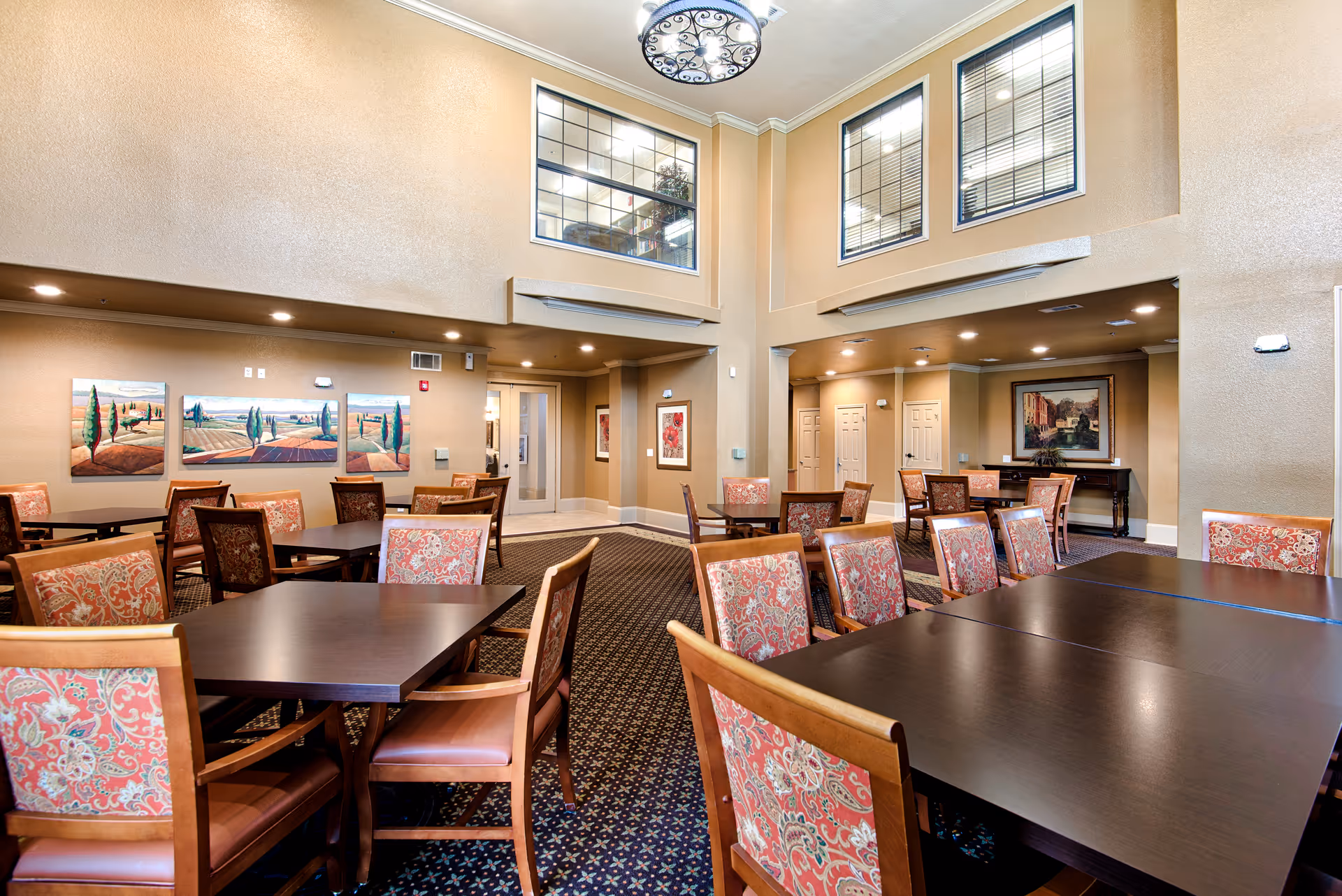 A spacious dining room with multiple dark wood tables and chairs featuring patterned upholstery. The room has high ceilings with large windows near the ceiling, beige walls, and decorative paintings on the walls. The carpet has a dark pattern, and there is a chandelier hanging from the ceiling.